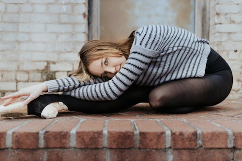 Full body slim agile female dancer in pointe shoes lying with arms crossed on leg on brick floor outside shabby building and looking at camera