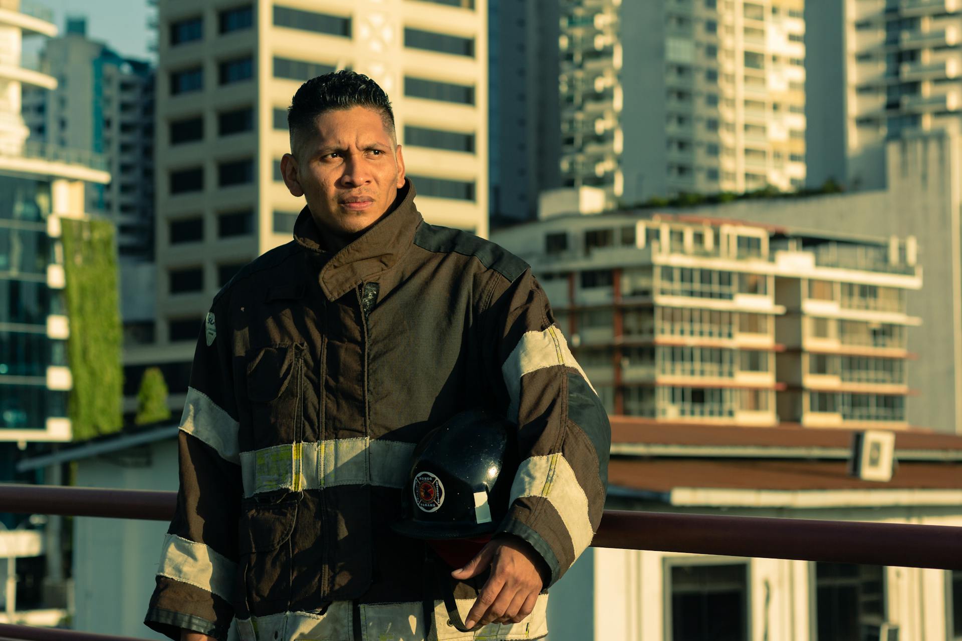 Firefighter standing outdoors in protective gear holding helmet, urban background.