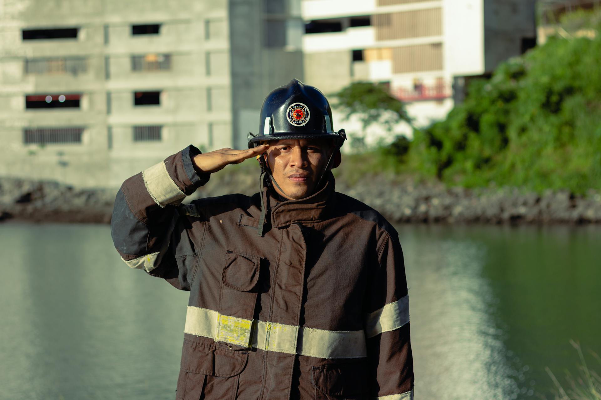 Firefighter in uniform saluting near a waterfront with buildings in the background.