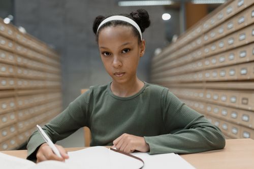 Free A young girl with a serious expression writing in a library with card catalogs. Stock Photo