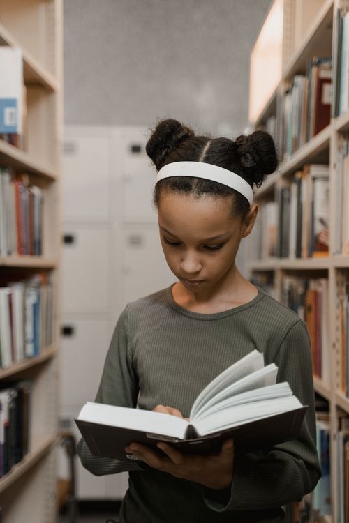 Free A young girl standing in a library aisle, reading a book with focused attention. Stock Photo