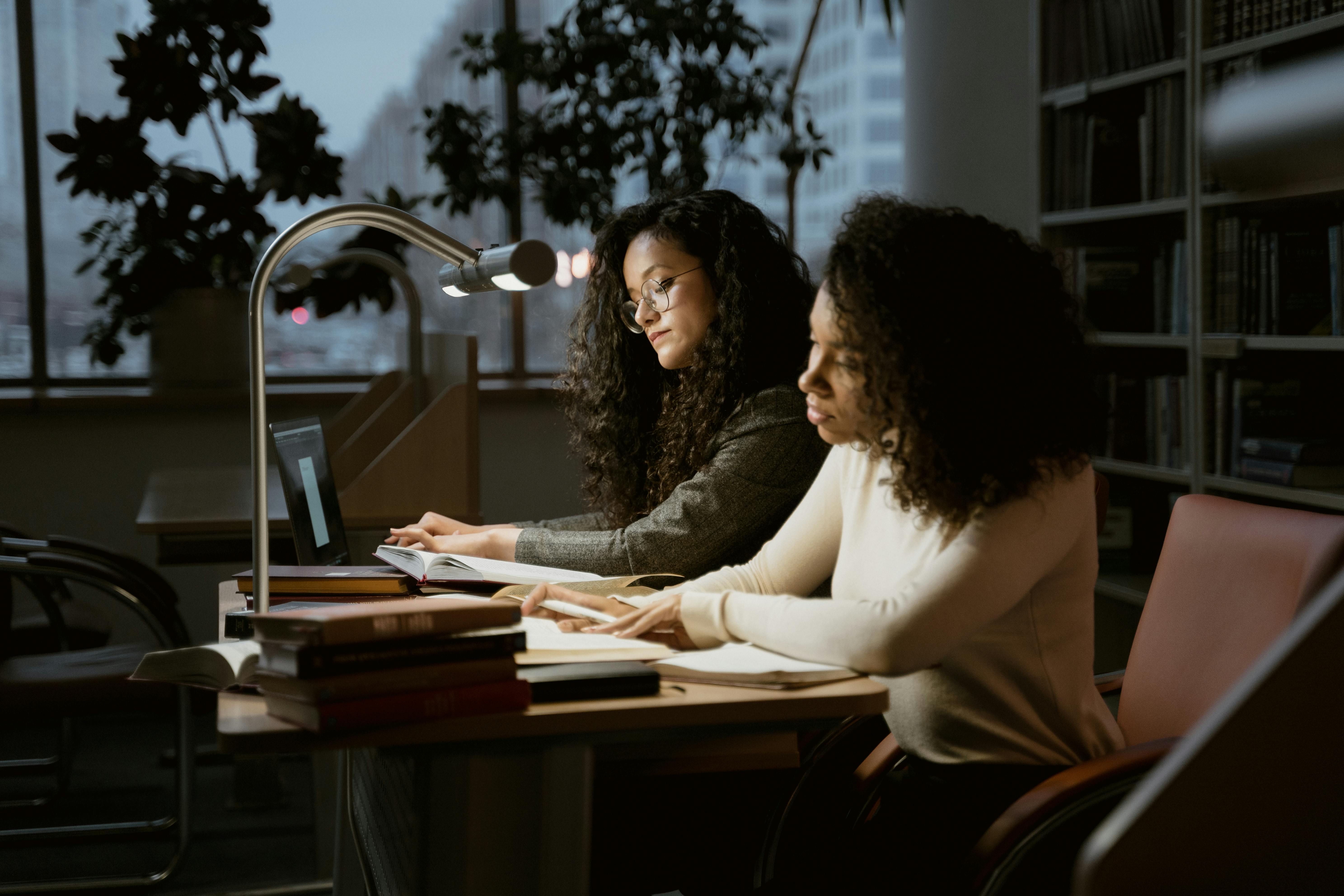 Two women intensely focused on studying in a library setting with laptops and books.