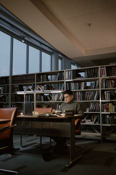 A student deeply engaged in study at a library desk surrounded by bookshelves and natural light.