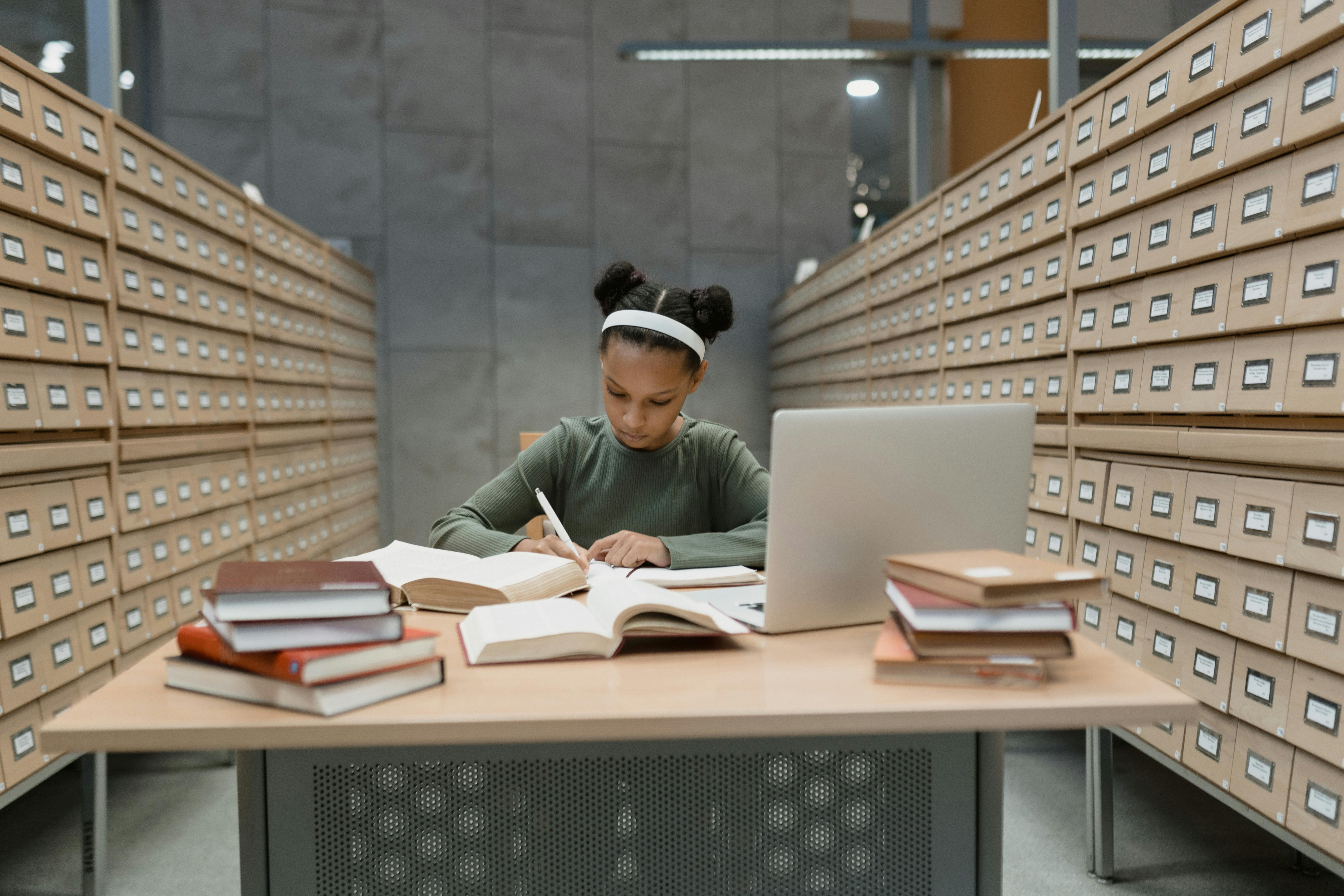 Young girl studying at a library table with books and laptop, deep in concentration.