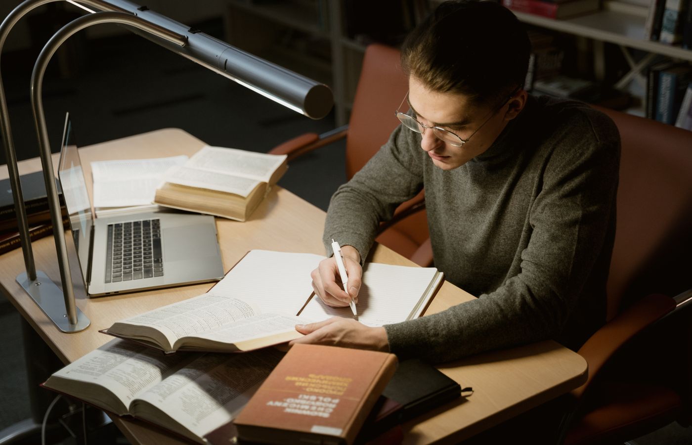 A young adult studying with books and a laptop, focused on taking notes at a desk under a lamp.