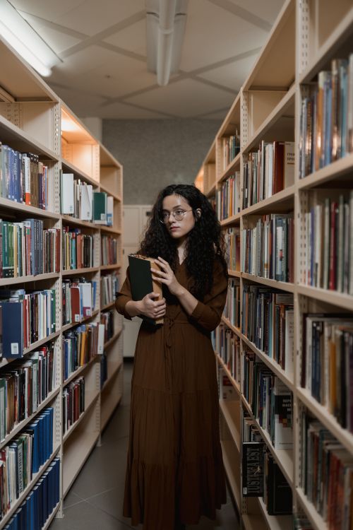 Free A young woman holding a book stands between bookshelves in a library, deep in thought. Stock Photo