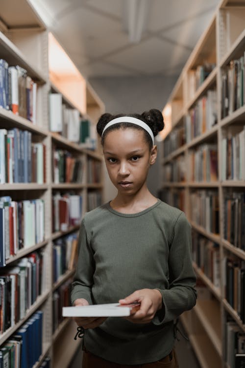 Free A Girl Holding a Book while Standing in a Library Stock Photo