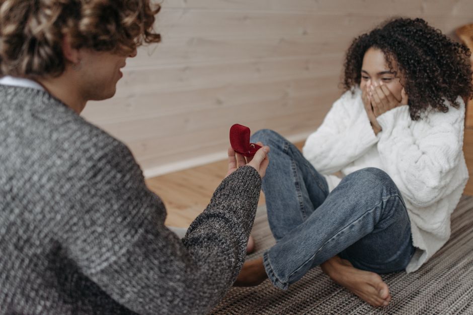 A man proposing to a woman indoors with a red ring box, capturing a moment of surprise and joy.