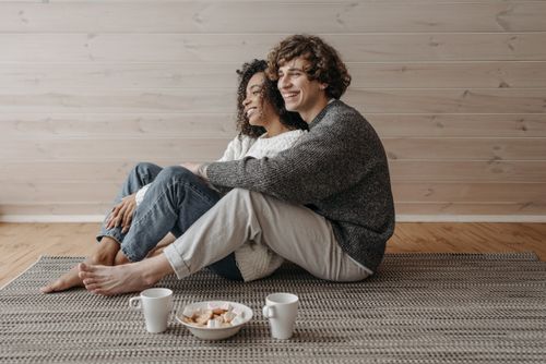 Free A happy couple sitting indoors on a carpet, barefoot and smiling, enjoying a cozy moment together. Stock Photo
