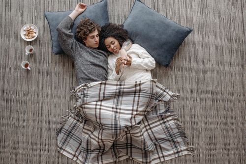 Free Interracial couple cuddling on the floor under a plaid blanket, enjoying a warm and cozy moment. Stock Photo