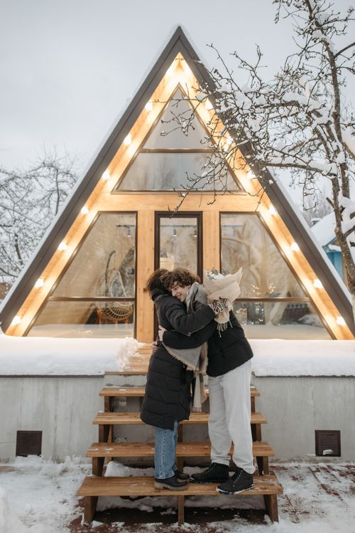 Free Couple embracing outside a triangular A-frame cabin in a snowy winter scene. Stock Photo