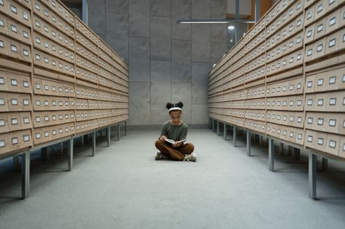 Free A girl sits reading a book in a library archive with wooden drawers. Stock Photo