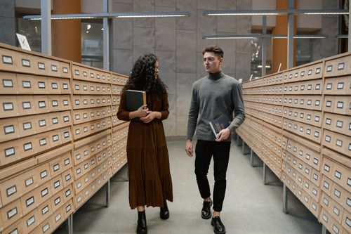 Free Two students walk through a library archive, each holding books and conversing. Stock Photo