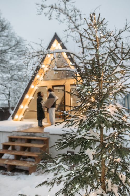 Free A serene winter scene featuring a lit A-frame cabin amidst snowy trees and festive lights. Stock Photo