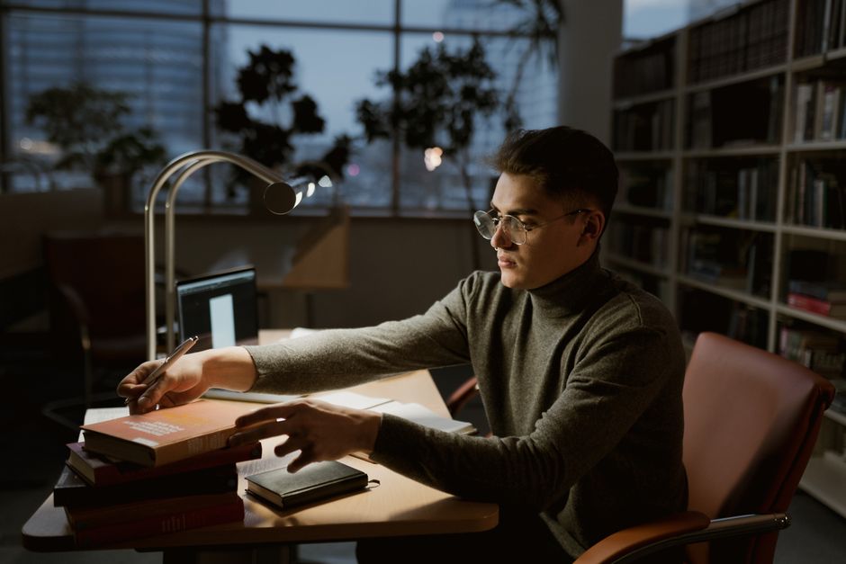 A young man wearing a turtleneck studies at a desk in a dimly lit library.