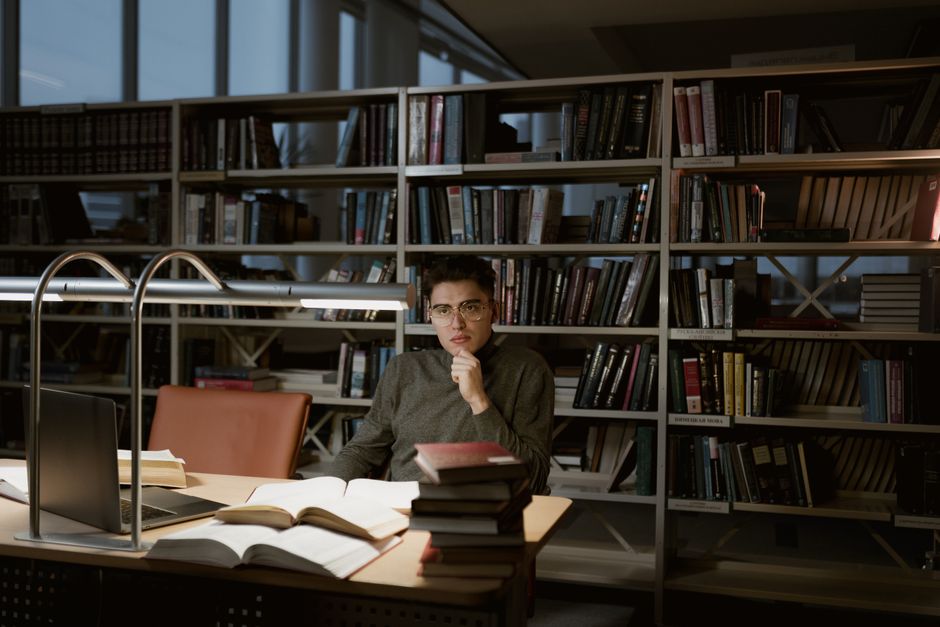 Young man sitting in library, studying with books, glasses, and laptop. Young man sitting in library, studying with books, glasses, and laptop.