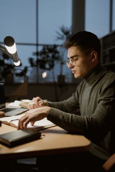 A young man in glasses studying at a desk with a book and notes in a cozy indoor setting.