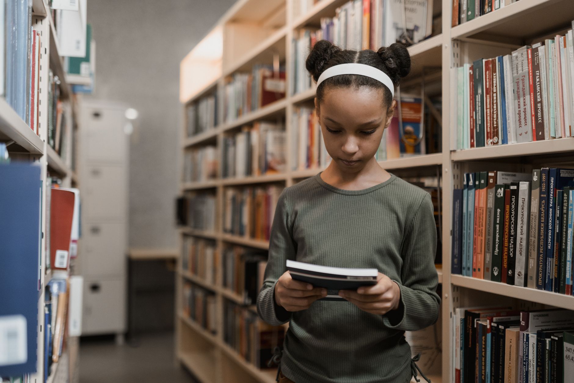 A young girl engrossed in reading a book while standing in a quiet library aisle.
