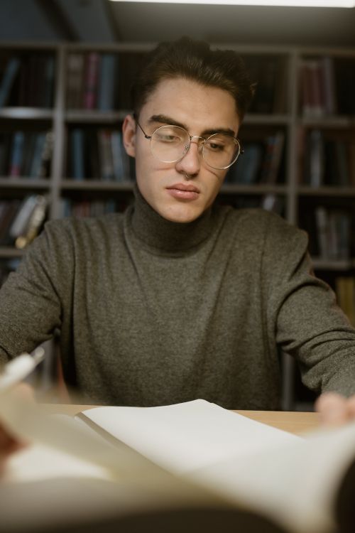 Free Focused young man with glasses studying in a library environment. Stock Photo