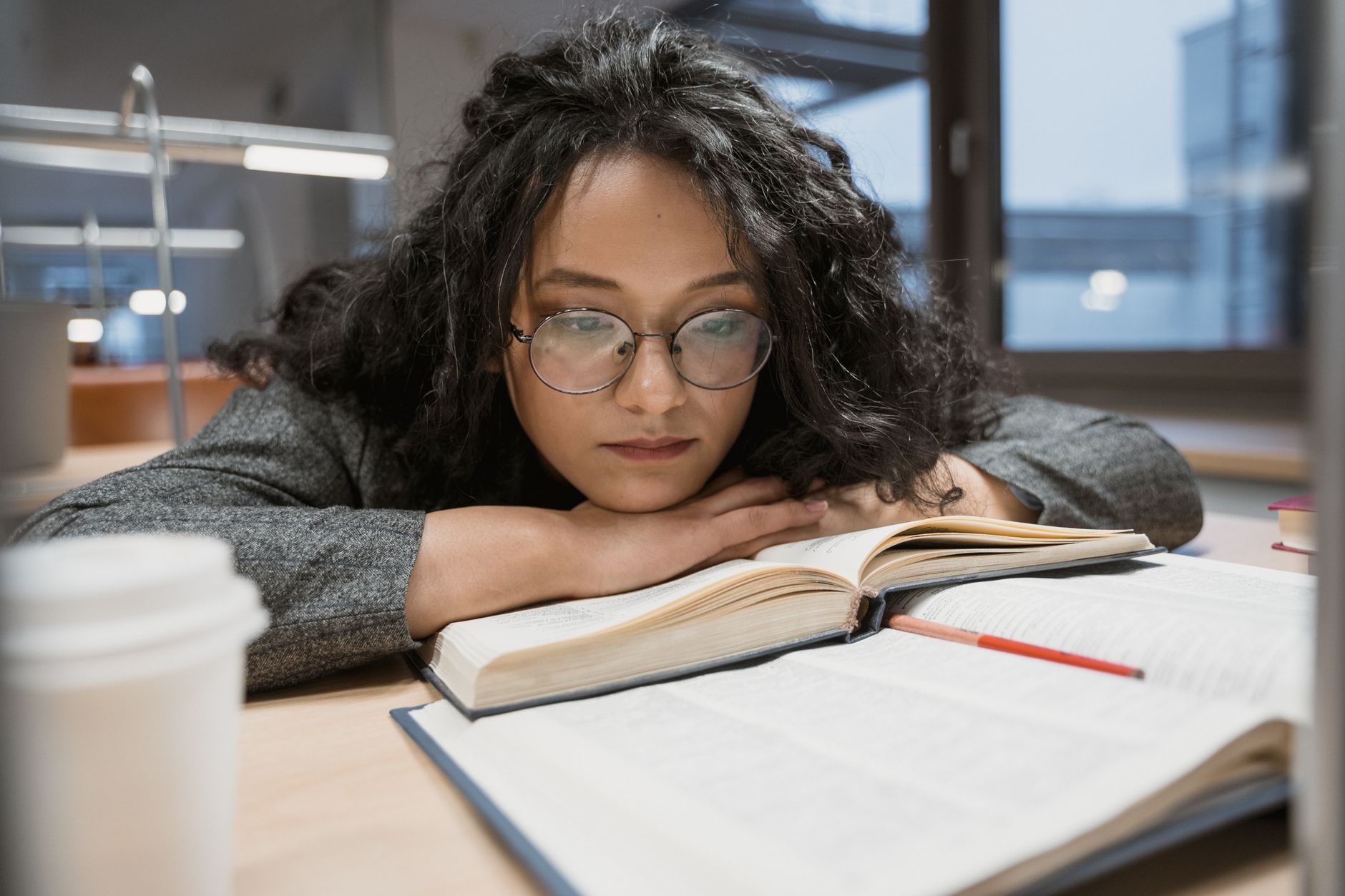 A woman with glasses deeply immersed in study, surrounded by books and a coffee cup in a library.
