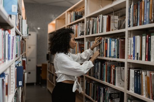 Free A young woman with curly hair searches for books on a library bookshelf. Stock Photo