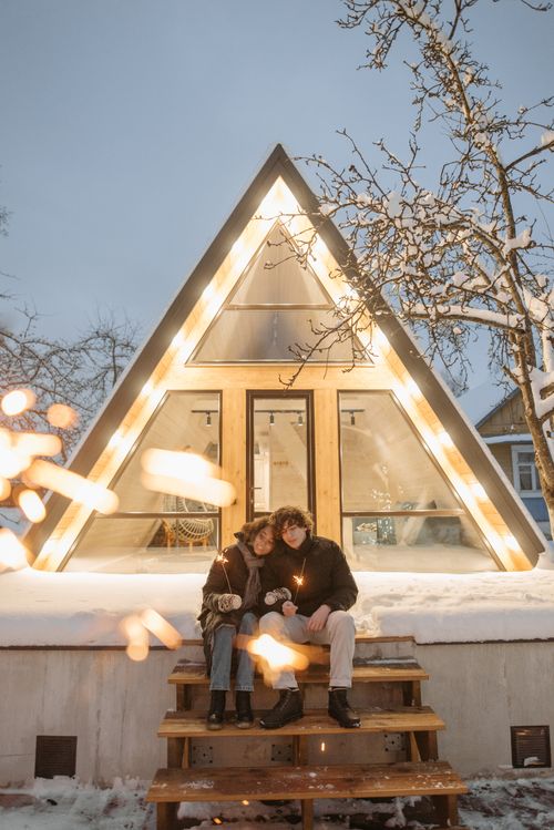 Free A romantic couple holds sparklers in front of a snow-covered A-frame cabin, capturing a cozy winter moment. Stock Photo
