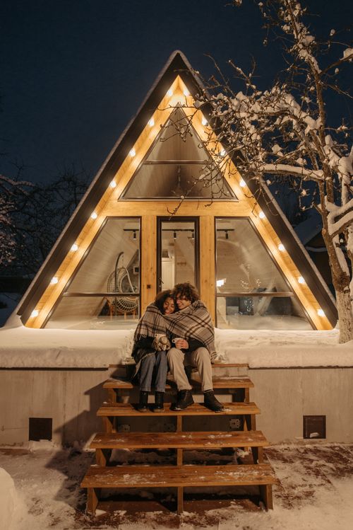 Free A couple sits warmly wrapped on steps of an A-frame cabin on a snowy winter night, enjoying a romantic moment. Stock Photo