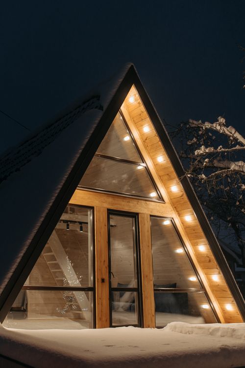 Free Warmly lit A-frame house with snow, triangular facade and glass windows at night. Stock Photo