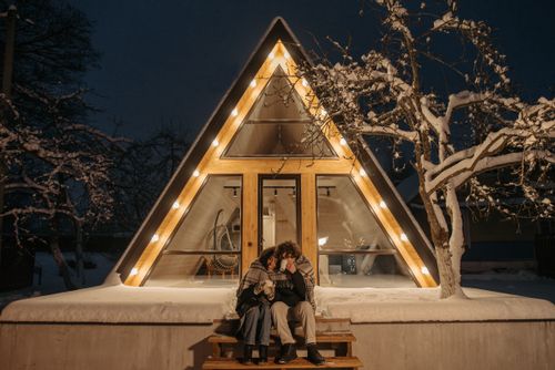 Free A couple sits warmly on stairs of a snow-covered A-frame cabin, sharing hot drinks in winter's embrace. Stock Photo