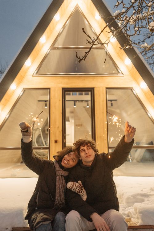 Free A couple sitting outside a triangular cabin, enjoying sparklers and winter ambiance. Stock Photo