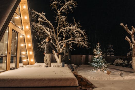 Couple walking hand in hand by an illuminated A-frame house on a snowy winter night.