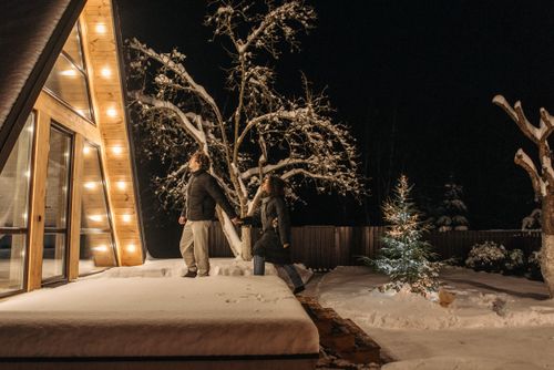 Free Couple walking hand in hand by an illuminated A-frame house on a snowy winter night. Stock Photo