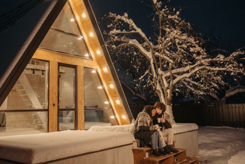 Free Couple enjoying a warm embrace outside an illuminated A-frame cabin on a snowy winter night. Stock Photo