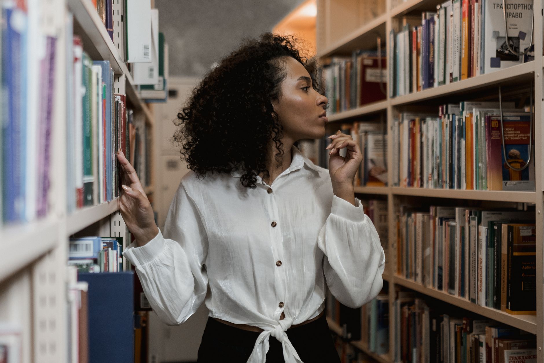 Curly-haired woman browsing bookshelves in a library, standing thoughtfully.
