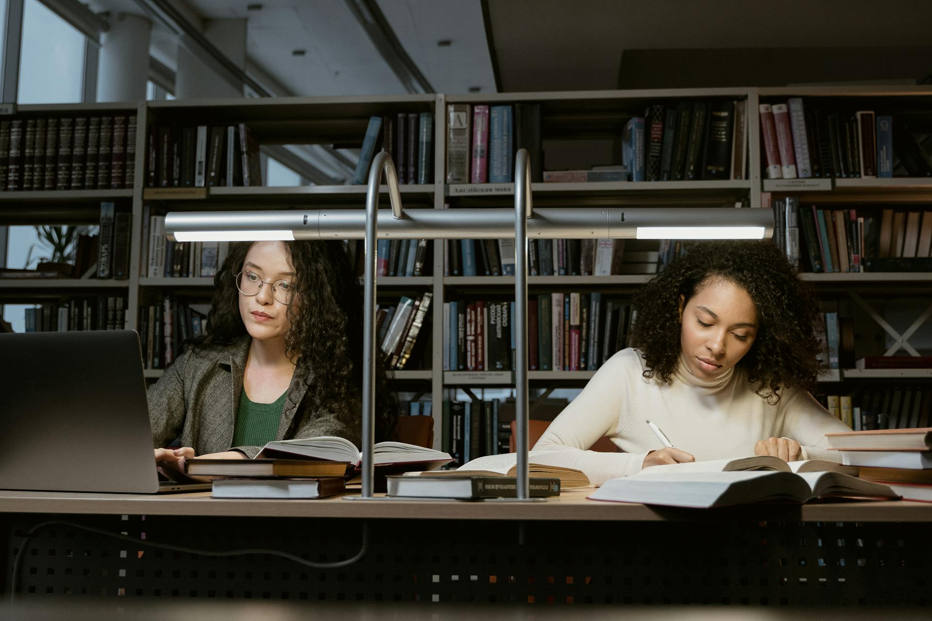 Two female students studying with books and laptops in a modern library setting.