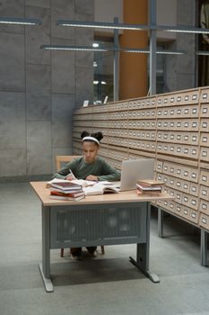 Focused girl studying at a library desk with books and laptop, deep in research.