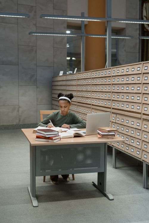 Free Focused girl studying at a library desk with books and laptop, deep in research. Stock Photo