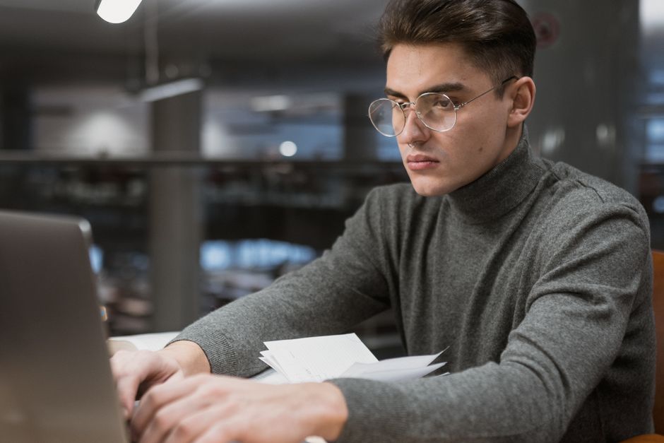 現代の大学図書館でノートパソコンを使って勉強する眼鏡をかけた若い男性。