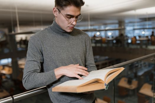 Adult man in gray turtleneck reading a book inside a modern library.