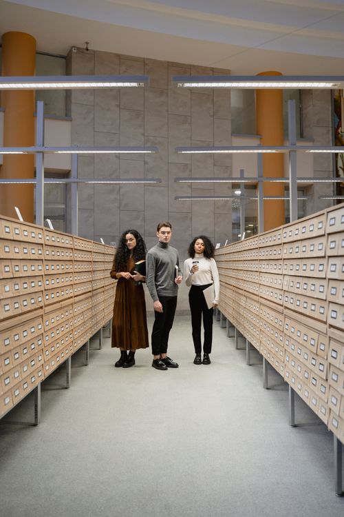 Free Three students browsing a card catalog aisle in a modern library setting. Stock Photo