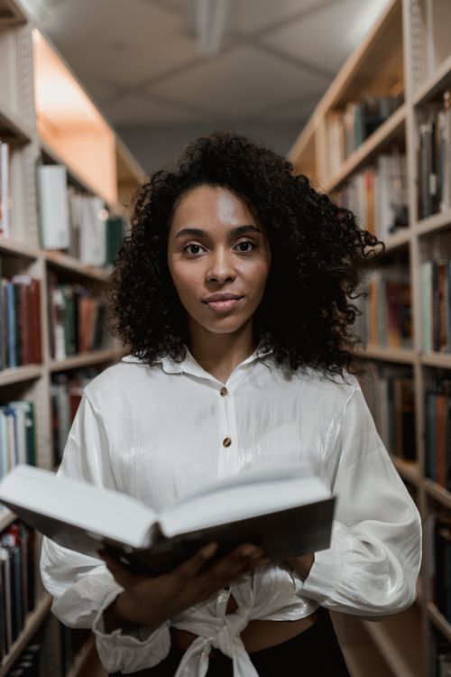 Free Portrait of a young woman with curly hair holding a book in a library setting. Stock Photo