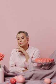 Sophisticated woman in soft pink attire sits beside an elegant cake on stand. Pastel backdrop enhances the serene mood.