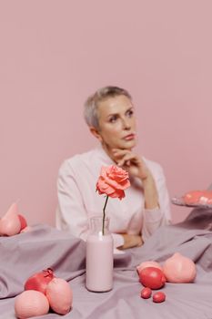 Thoughtful woman in pink with a rose, surrounded by pastel fabric and fruits on a pink backdrop.