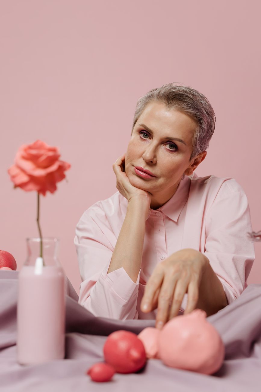 A mature woman poses thoughtfully among pink objects, creating a surreal aesthetic.