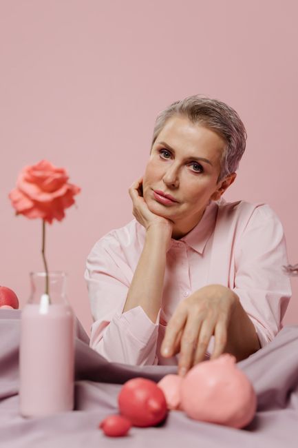 A mature woman poses thoughtfully among pink objects, creating a surreal aesthetic.