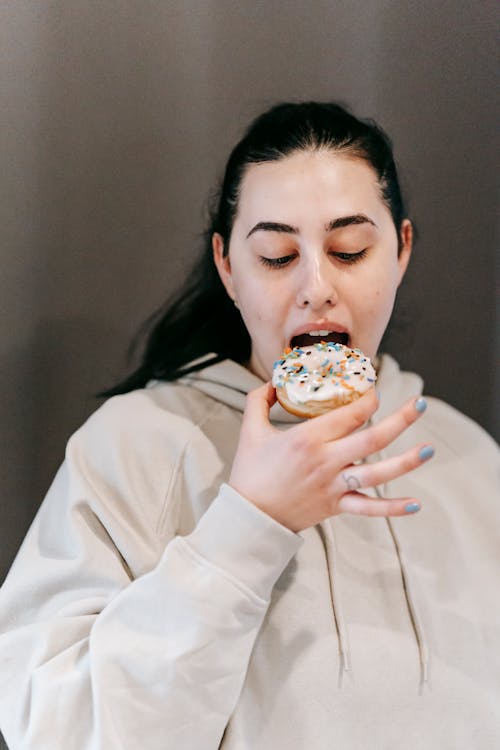Glad young female in casual clothes biting yummy doughnut with vanilla icing in light room