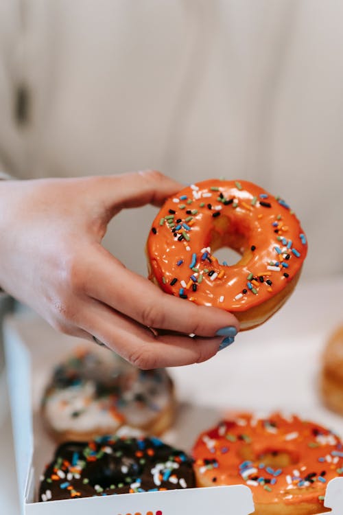 Crop anonymous female with manicured hands showing yummy glazed doughnut decorated with sprinkles in light room