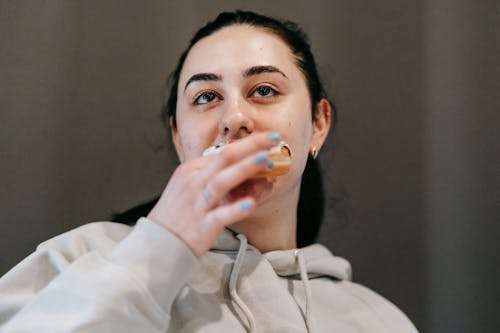 Glad young brunette in comfy hoodie biting delicious glazed doughnut and looking away in light room