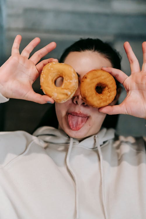 Cheerful grimacing female in loose clothes holding delicious doughnuts against eyes and showing tongue in light room