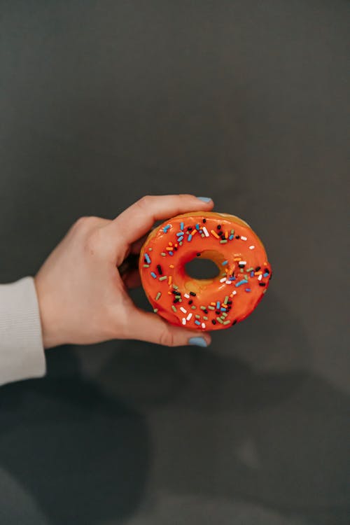 Crop unrecognizable female with manicured hands demonstrating delicious fresh glazed doughnut against dark gray wall